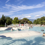 Menschen genießen einen sonnigen Tag in einem großen, flachen Pool mit Wasserspielen.