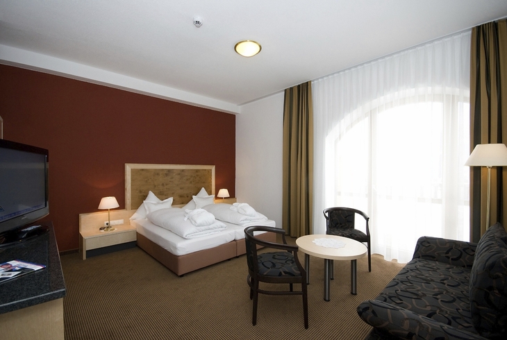 Hotelzimmer mit Doppelbett, Sofa, Tisch und Stühlen, Blick auf großes Fenster.