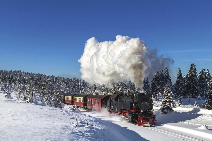 Dampflok fährt durch verschneite Landschaft mit Nadelbäumen unter klarem, blauem Himmel.