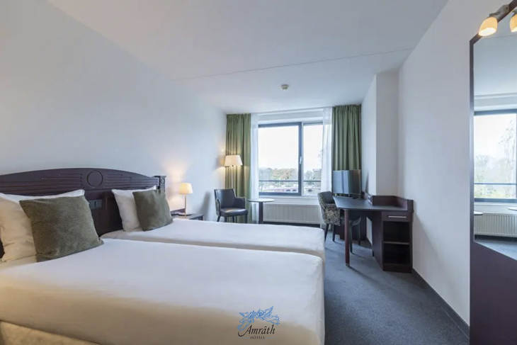 Hotelzimmer mit zwei Betten, Schreibtisch, Lampe und Fenster mit Vorhängen.