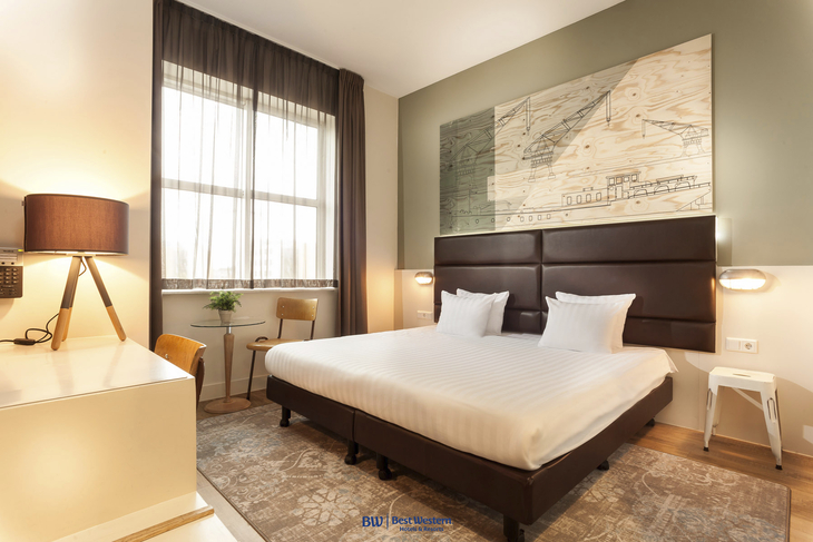 Hotelzimmer mit Doppelbett, Lampe, großem Fenster und Wandkunst in modernem Stil.