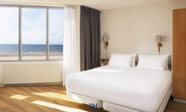 Helles Hotelzimmer mit großem Bett und Meerblick aus dem Fenster.