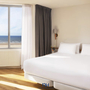 Helles Hotelzimmer mit großem Bett und Meerblick aus dem Fenster.