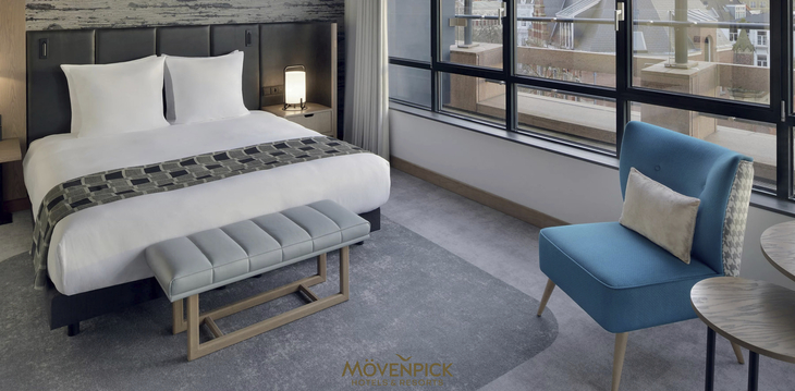 Innenansicht eines modernen Hotelzimmers mit Doppelbett und blauer Sitzgelegenheit.