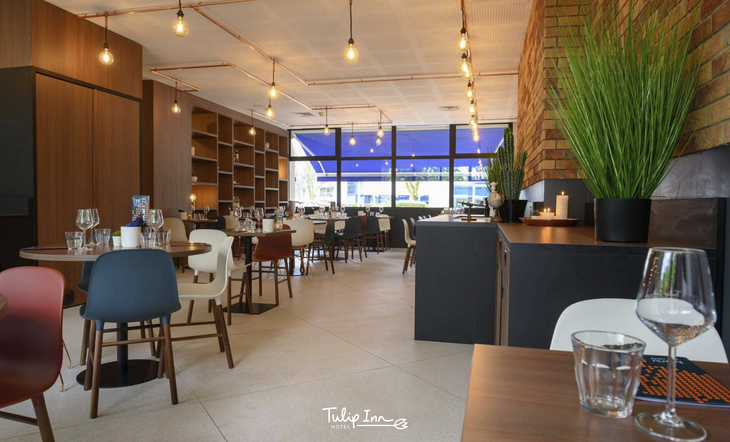 Modernes Hotelrestaurant mit gedeckten Tischen, Pflanzen und großen Fenstern.