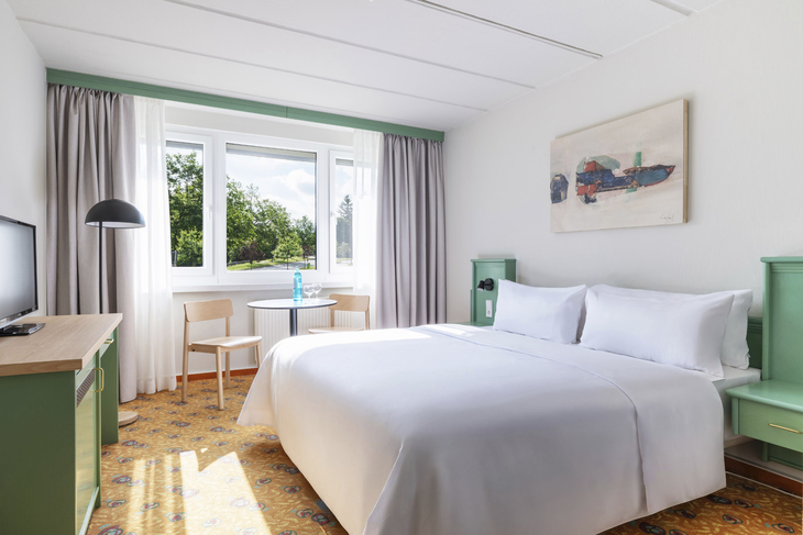 Helles Hotelzimmer mit Doppelbett, Tisch und großem Fenster mit Blick ins Grüne.