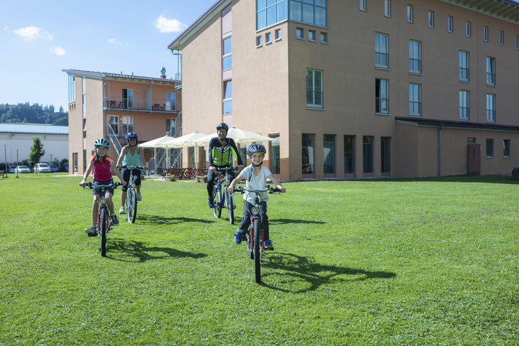 Menschen fahren Fahrrad vor JUFA Kempten Familien-Resort auf grüner Wiese.