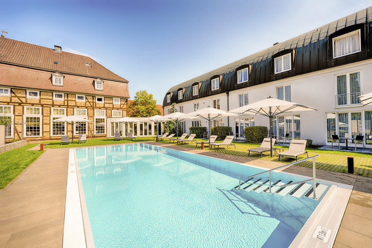 Hotelpool mit Liegestühlen und Sonnenschirmen, umgeben von Gebäuden bei sonnigem Wetter.