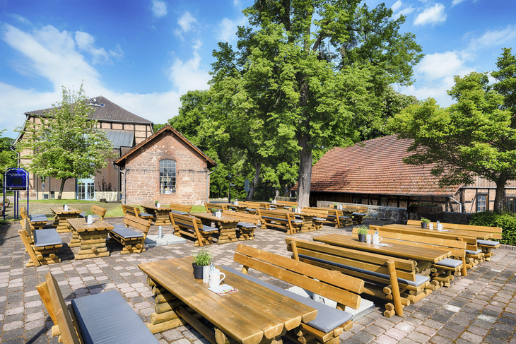 Biergarten mit Holzbänken und Tischen auf gepflastertem Platz, umgeben von Bäumen.
