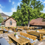Biergarten mit Holzbänken und Tischen auf gepflastertem Platz, umgeben von Bäumen.
