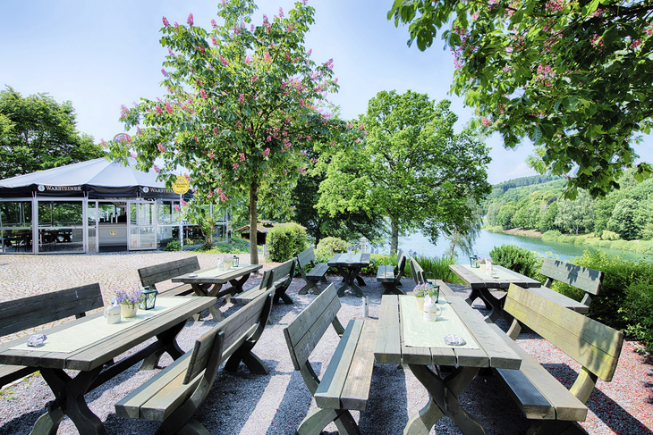 Biergarten mit Holztischen unter blühenden Bäumen am Ufer eines Sees.