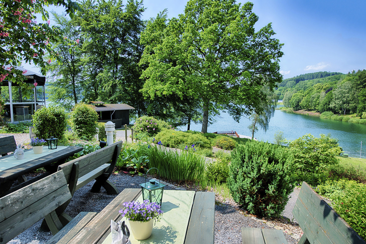 Biergarten mit Holzbänken, Aussicht auf einen See und grüne Bäume im Hintergrund.
