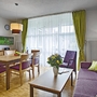 Wohnbereich der Ferienanlage Titisee mit Holzfußboden, Esstisch und Stühlen aus Holz, sowie einem lila Sofa und Sessel mit kleinem Holztisch davor, auf dem eine Vase mit Blumen steht. Links, vor dem großen Fenster mit weißer Gardine und grünem Vorhang, hängt ein Flachbildschirm an der Wand über einem Sideboard unter einer Wanduhr.