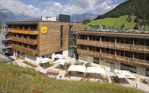 Hotelgebäude mit Terrasse und Bergkulisse an einem sonnigen Tag.