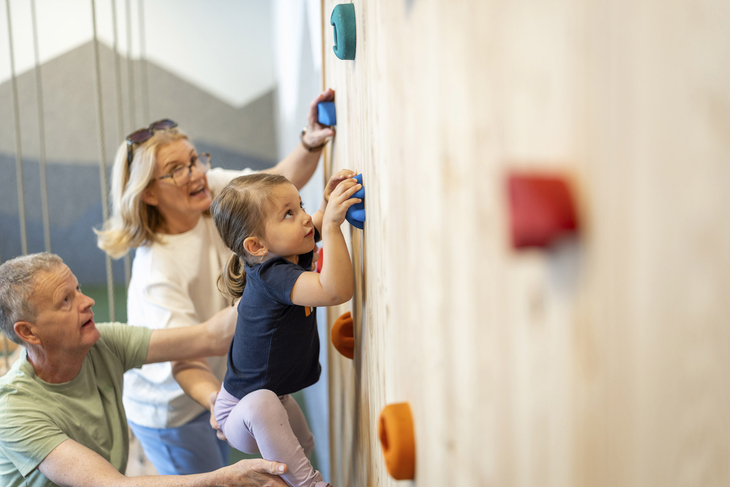 Kind klettert mit Hilfe von Erwachsenen an einer Indoor-Kletterwand.