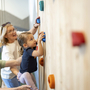 Kind klettert mit Hilfe von Erwachsenen an einer Indoor-Kletterwand.