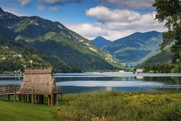 Blick auf den Ledrosee in Italien mit umliegenden Bergen und Schilf am Ufer.
