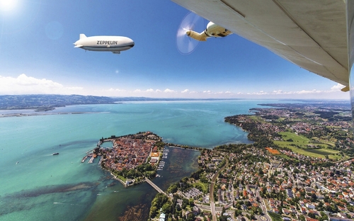Luftaufnahme eines Zeppelins über einer Stadt an einem großen See.