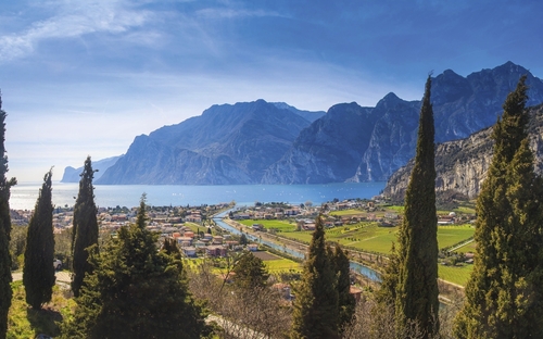 Blick auf den Gardasee mit Bergen im Hintergrund und Zypressen im Vordergrund.