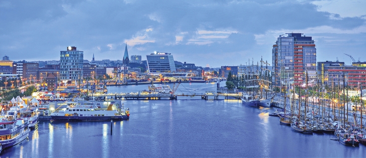 Blick auf den Hafen von Kiel bei Abenddämmerung mit beleuchteten Gebäuden und Segelschiffen.