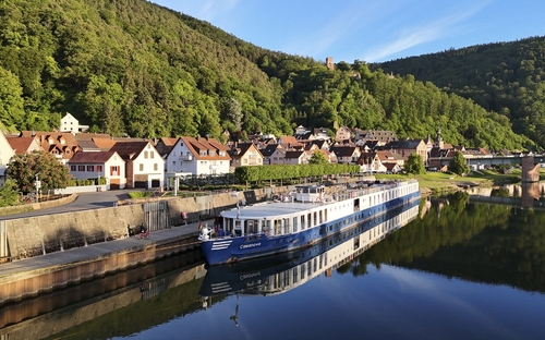 Flusskreuzfahrtschiff an einem sonnigen Tag vor einer bewaldeten Hügellandschaft und Dorf.
