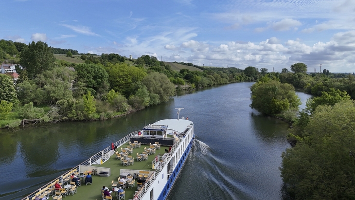 Flusskreuzfahrtschiff fährt auf einem von Bäumen gesäumten Fluss bei sonnigem Wetter.