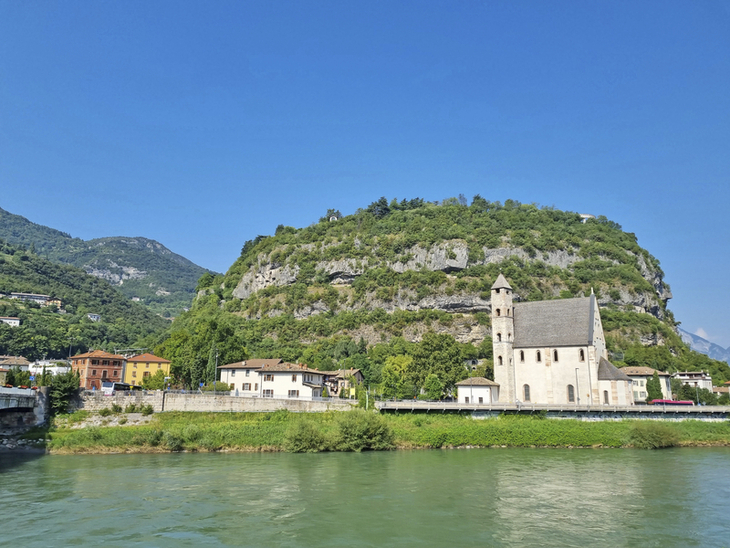 Kirche am Flussufer vor bewaldetem Hügel unter klarem Himmel.