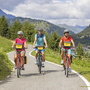 Drei Radfahrer auf einem Bergweg mit Alpen im Hintergrund.