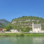 Kirche am Fluss vor bewaldetem Hügel und blauem Himmel.