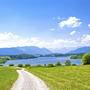 Landschaft mit Schotterweg, Wiesen, einem See und Bergen im Hintergrund bei sonnigem Wetter.