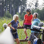 Drei Radfahrer machen Pause am Seeufer in einer grünen Landschaft.