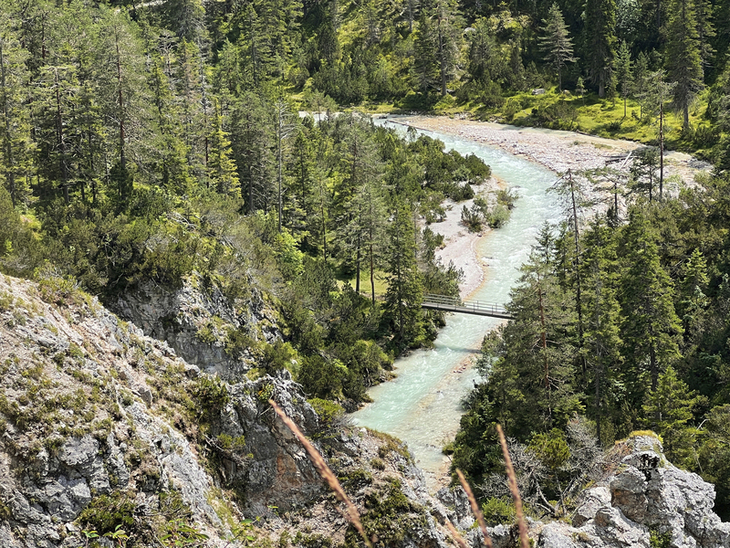 Fluss in einer alpinen Landschaft mit Brücke und Wald im Hintergrund.
