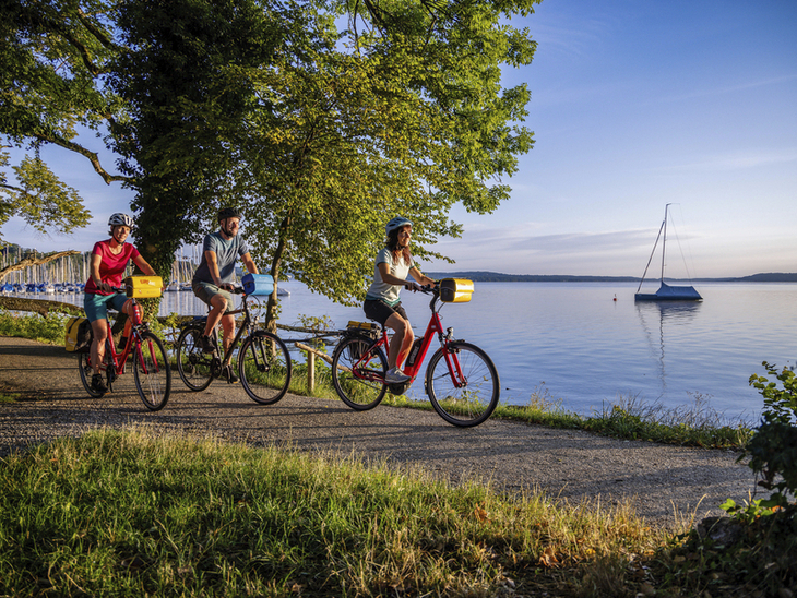 Radfahrer fahren am Starnberger See entlang, im Hintergrund Boote im Hafen.