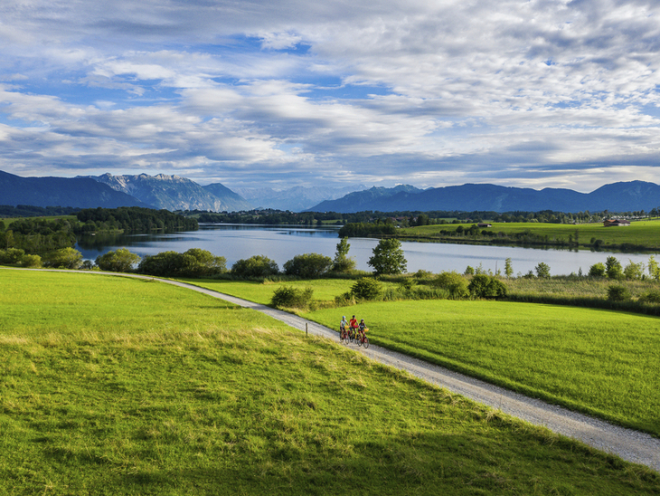 Radfahrer auf einem Weg am Riegsee vor Bergkulisse und Wiesenlandschaft.