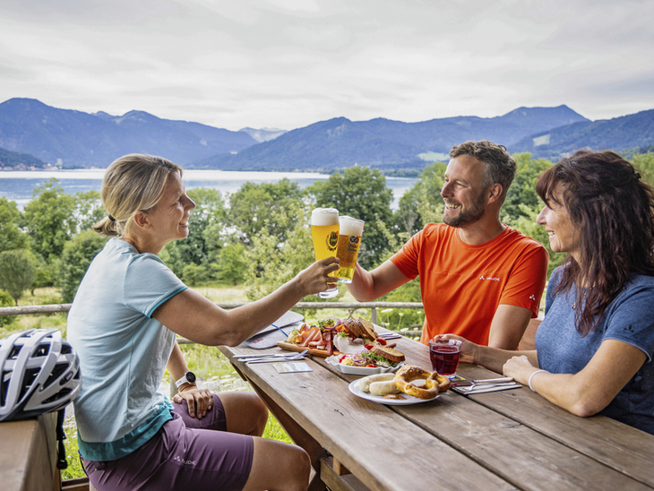 Drei Personen stoßen bei Brotzeit mit Bier am Tegernsee an.