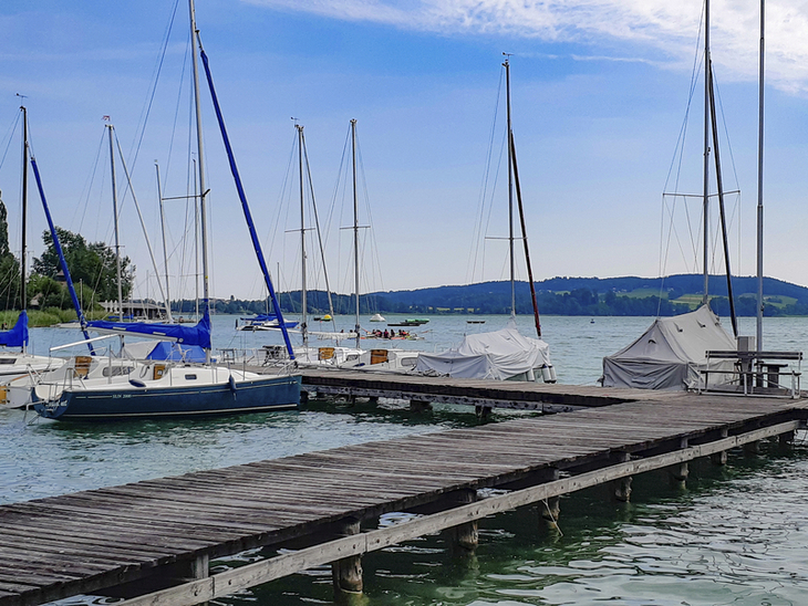 Segelboote an einem Steg auf einem ruhigen See mit Hügeln im Hintergrund