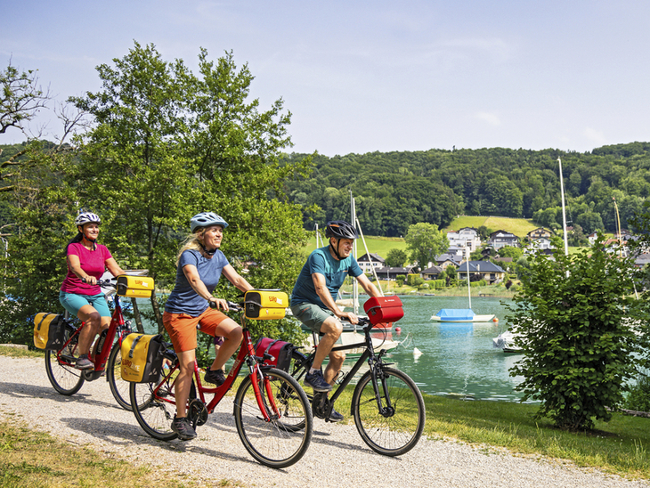 Drei Radfahrer fahren auf einem Weg entlang eines Sees bei schönem Wetter.