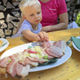 Kind greift nach Speckbrot an einem Holztisch im Freien.