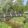 Radfahrer überqueren eine Holzbrücke in einer grünen, natürlichen Umgebung.