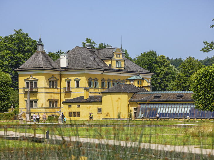 Gelbes Schloss Hellbrunn in Salzburg mit modernem Anbau und Garten im Vordergrund.