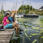 Drei Frauen sitzen auf einem Steg am See, Boote und Ufer im Hintergrund.
