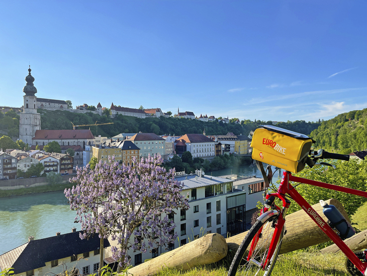 Fahrrad vor Blick auf Burghausen mit Fluss und blühendem Baum im Vordergrund.