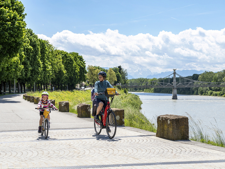 Familie beim Radfahren entlang eines Flusses mit Brücke im Hintergrund.