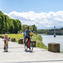 Familie beim Radfahren entlang eines Flusses mit Brücke im Hintergrund.