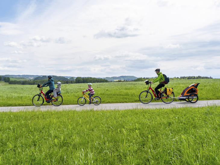 Familie radelt auf einem Weg durch eine grüne Landschaft, Kinderanhänger dabei.