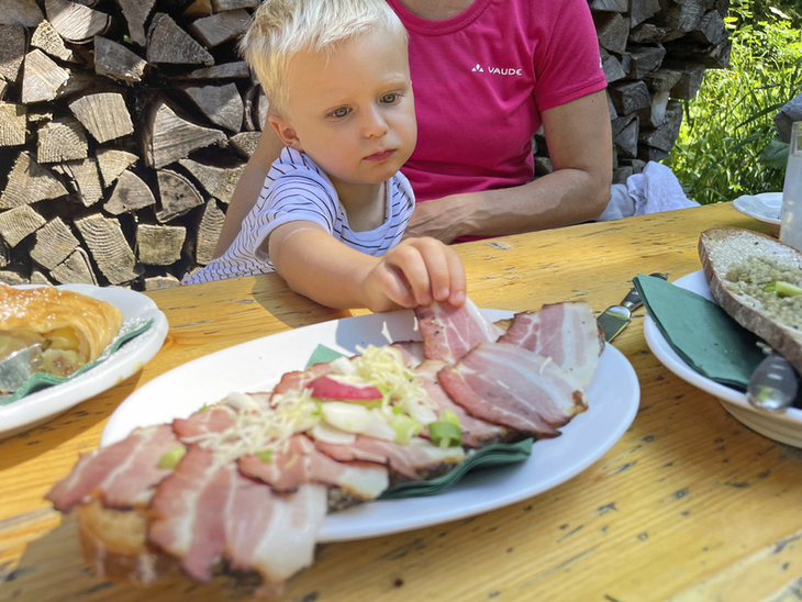 Kind greift nach Speckbrot auf Holztisch im Freien.