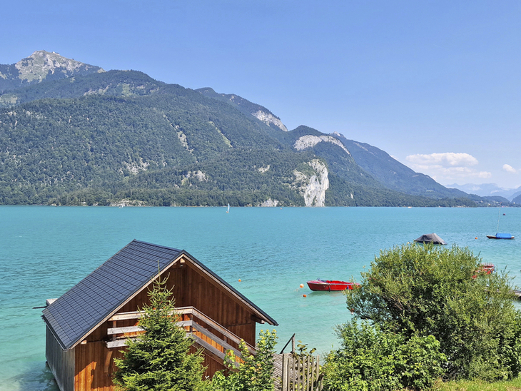 Holzhütte am Wolfgangsee mit Bergen im Hintergrund und blauem Himmel.