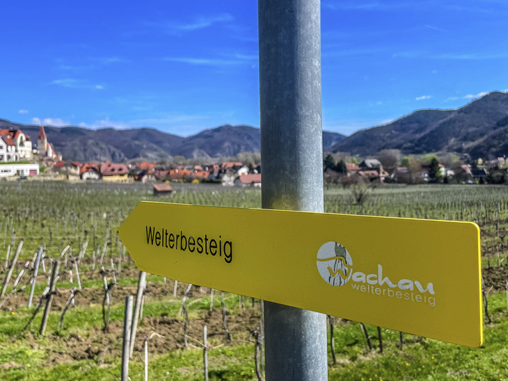Wegweiser zum Welterbesteig Wachau vor Weinbergen und Dorf in der Ferne.