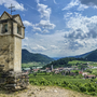 Kleines Denkmal am Hang, Blick auf grünes Tal mit Dorf und Bergen im Hintergrund.