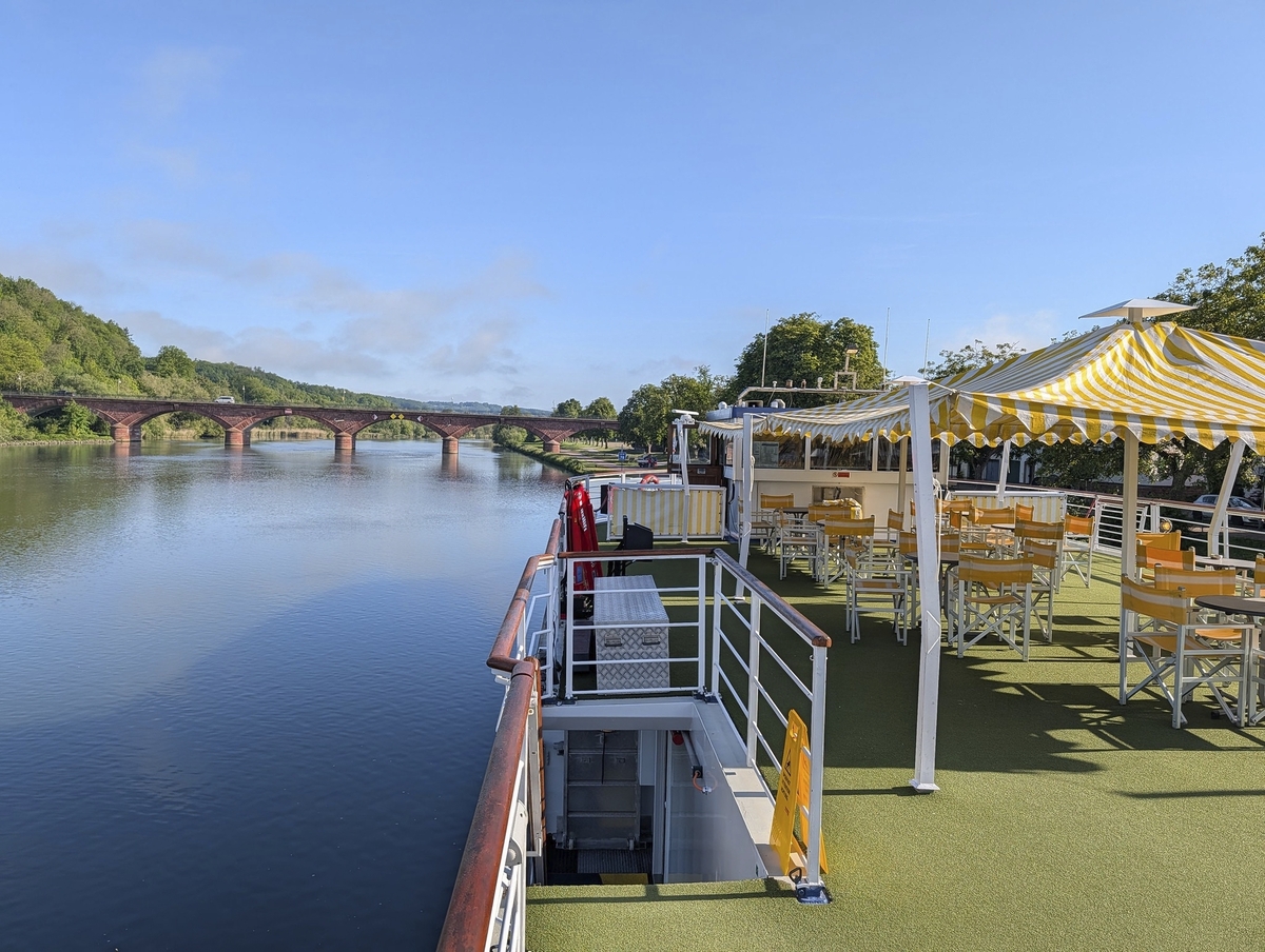 Sonnendeck eines Flusskreuzfahrtschiffs mit Tischen und Stühlen, Blick auf Brücke und Fluss.